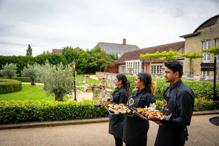 Two servers carrying canapés through landscaped grounds