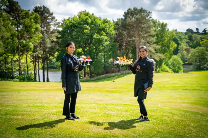 Two servers carrying trays across a sunny lawn