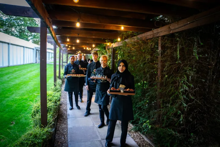 Servers carrying canapés through a garden passageway